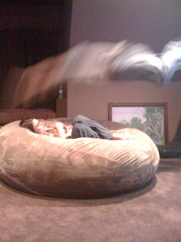 A picture of a husband leaping onto an unsuspecting wife who is curled up in a brown, giant Fombag watching TV