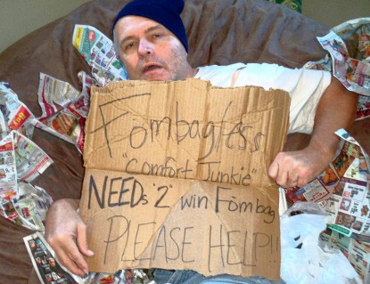 A picture of a man pretending to be a 'comfort junkie' holding up a sign as he lays on a brown, giant Fombag